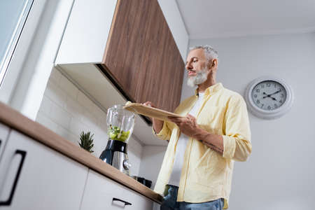 Low angle view of tattooed man holding cutting board near blender with fruits in kitchenの写真素材