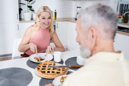 Smiling woman cutting pie near blurred husband in kitchenの写真素材