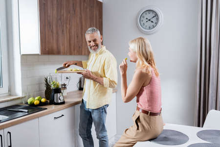 Positive man adding fruits in blender near wife in kitchenの写真素材