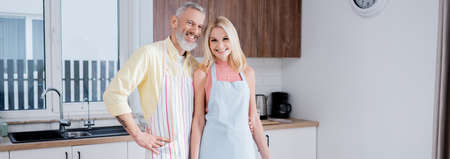 Positive mature couple in aprons looking at camera in kitchen, bannerの写真素材