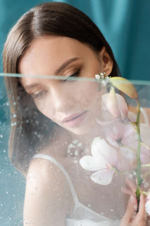 young and sensual woman with white orchid near blurred wet glass on turquoise backgroundの写真素材