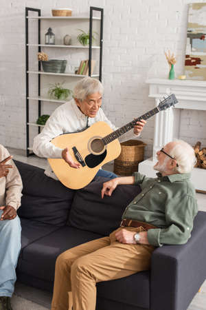 Senior asian man holding acoustic guitar while talking to friend on couch at homeの写真素材