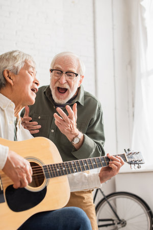 Excited senior man singing near asian friend with acoustic guitar at homeの写真素材