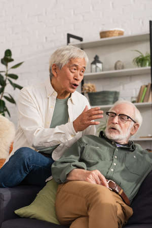 Elderly asian man talking near friend on couch at homeの写真素材