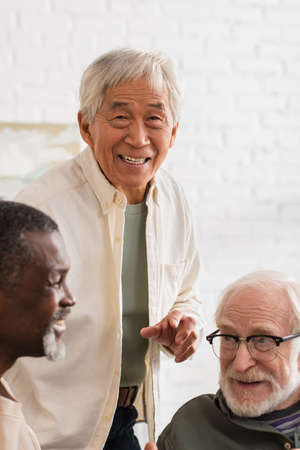 Happy asian man looking at camera near multiethnic friends at homeの写真素材