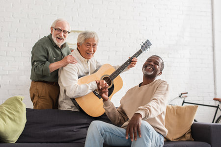 Happy senior interracial men with acoustic guitar looking at camera at homeの写真素材
