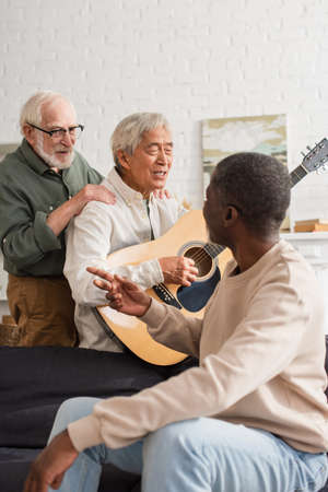 Positive elderly man hugging asian friend playing acoustic guitar in living roomの写真素材