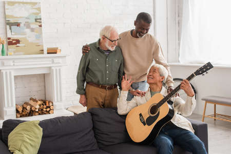 Smiling multiethnic men hugging asian friend with acoustic guitar at homeの写真素材