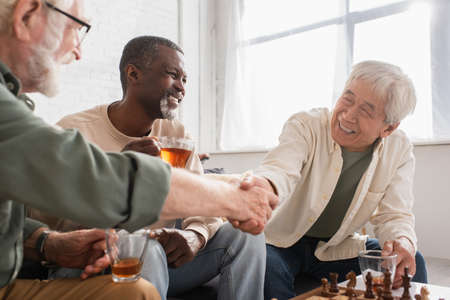 Interracial pensioners shaking hands near chessboard and african american friend with tea at home