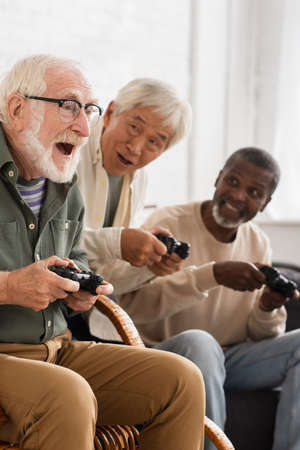 KYIV, UKRAINE - OCTOBER 12, 2021: Excited man playing video game with senior interracial friends at homeの写真素材