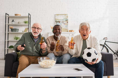 Multicultural senior friends with football and beer watching tv at homeの写真素材