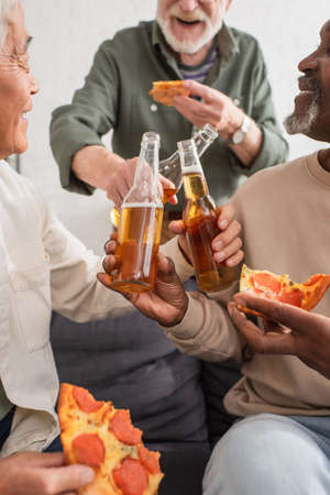 Interracial senior friends holding beer bottles and pizza at home