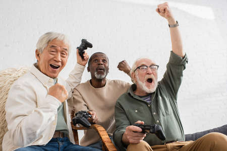 KYIV, UKRAINE - OCTOBER 12, 2021: Excited multicultural friends showing yes gesture while playing video game at homeのeditorial素材