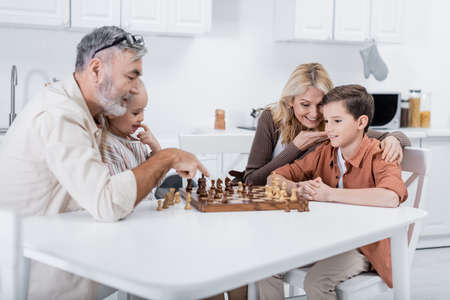 senior man pointing at chessboard near grandchildren and wife in kitchenの写真素材