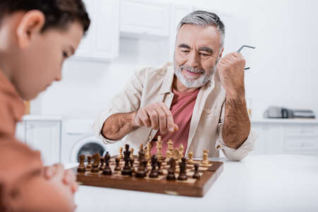 pleased senior man playing chess with blurred grandson in kitchenの写真素材
