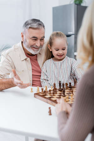 smiling senior man pointing with hand at chessboard near granddaughter and blurred wifeの写真素材