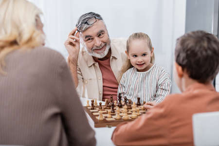smiling man holding eyeglasses while playing chess with wife and grandchildrenの写真素材