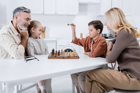 boy holding chess figure near smiling grandparents and sister in kitchenの写真素材