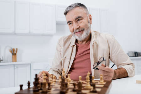 pleased grey-haired man holding eyeglasses while playing chess in kitchenの写真素材