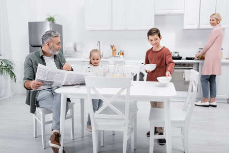 senior man with newspaper looking at granddaughter holding cups in kitchenの写真素材
