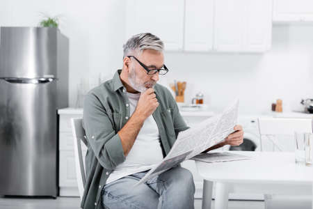 thoughtful senior man in eyeglasses reading morning newspaper in kitchenの写真素材
