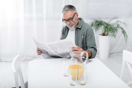 senior man in eyeglasses reading morning newspaper near jug with orange juiceの写真素材