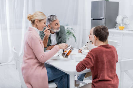 smiling middle aged woman pouring tea near grandchildren and husband during breakfast