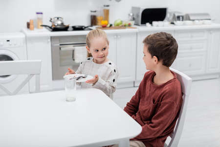 smiling girl holding plate with cutlery near brother sitting at kitchen tableの写真素材