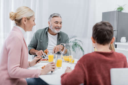 cheerful senior man having breakfast with blurred grandchildren and wife in kitchen