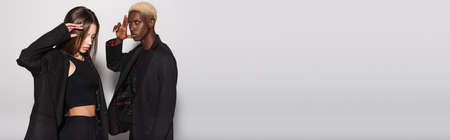 fashionable interracial couple in black suits posing on gray background with copy space, banner