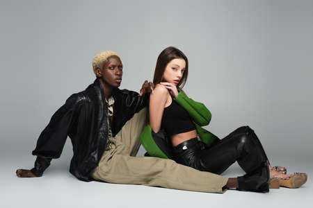 blonde african ameican man and sensual brunette woman sitting and looking at camera on gray