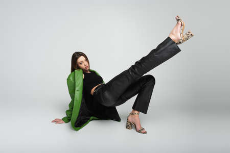 stylish woman in leather clothes and sandals sitting with raised leg on gray