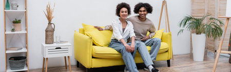 Cheerful african american couple looking at camera on couch near easel at home, bannerの写真素材