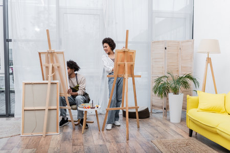 Smiling african american woman looking at boyfriend near canvases on easels at homeの写真素材