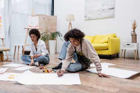 Positive african american man painting on paper on floor near girlfriend at homeの写真素材