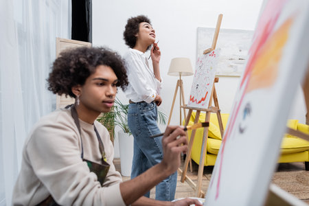 Dreamy african american woman holding paintbrush near canvas and blurred boyfriend painting at homeの写真素材