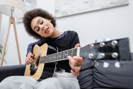 Young african american woman playing acoustic guitar on couch in living room
