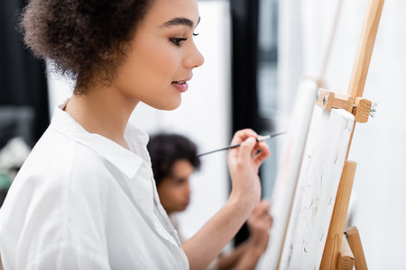 Side view of smiling african american woman holding paintbrush near canvas at home