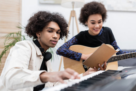 Smiling african american woman playing acoustic guitar near boyfriend with synthesizer and notebook at homeの写真素材