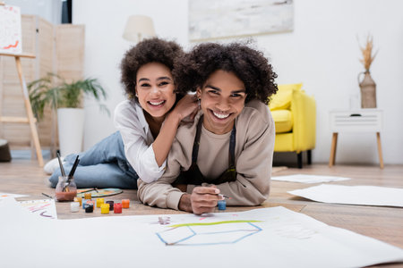 Happy african american woman hugging boyfriend and looking at camera near papers and paints on floorの写真素材