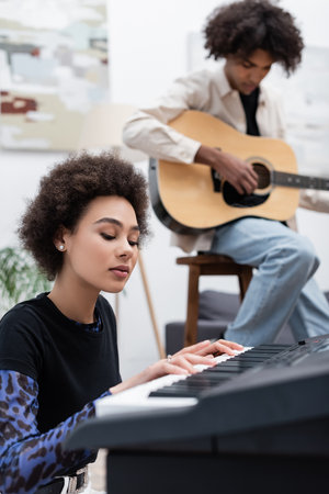 Young african american musician playing synthesizer near blurred boyfriend with acoustic guitar at homeの写真素材