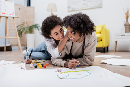 Positive african american woman hugging boyfriend with paintbrush near paper on floorの写真素材