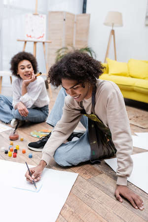 Smiling african american man painting on floor near blurred girlfriend at home