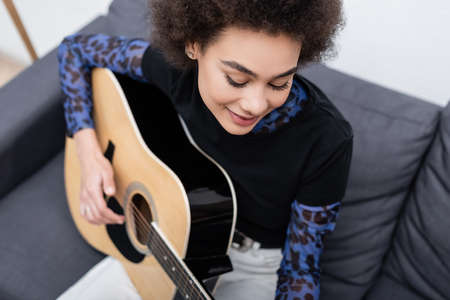 High angle view of smiling african american woman playing blurred acoustic guitar at home