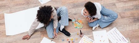 Overhead view of cheerful african american woman looking at boyfriend painting on floor at home, bannerの写真素材
