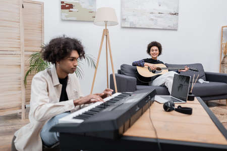 African american woman playing acoustic guitar near blurred boyfriend with synthesizer and laptop at homeの写真素材