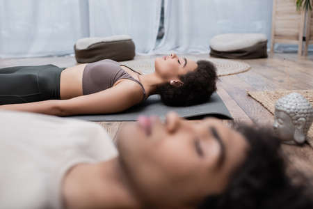 Young african american woman in sportswear meditating near blurred boyfriend at home
