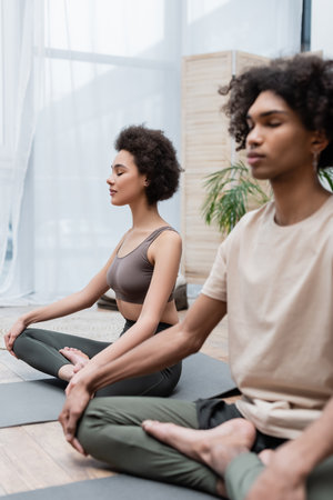 African american woman sitting in yoga pose near blurred boyfriend at homeの写真素材