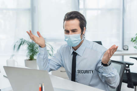 discouraged security man in medical mask gesturing near blurred laptop