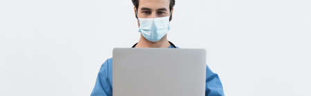 young doctor in medical mask using laptop isolated on grey, banner